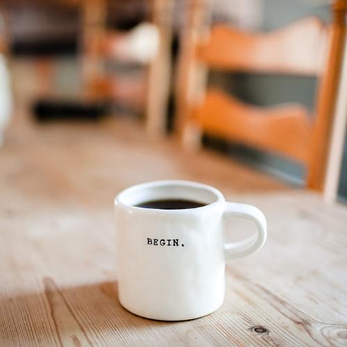 Begin printed on white coffee mug sitting on a wood table.